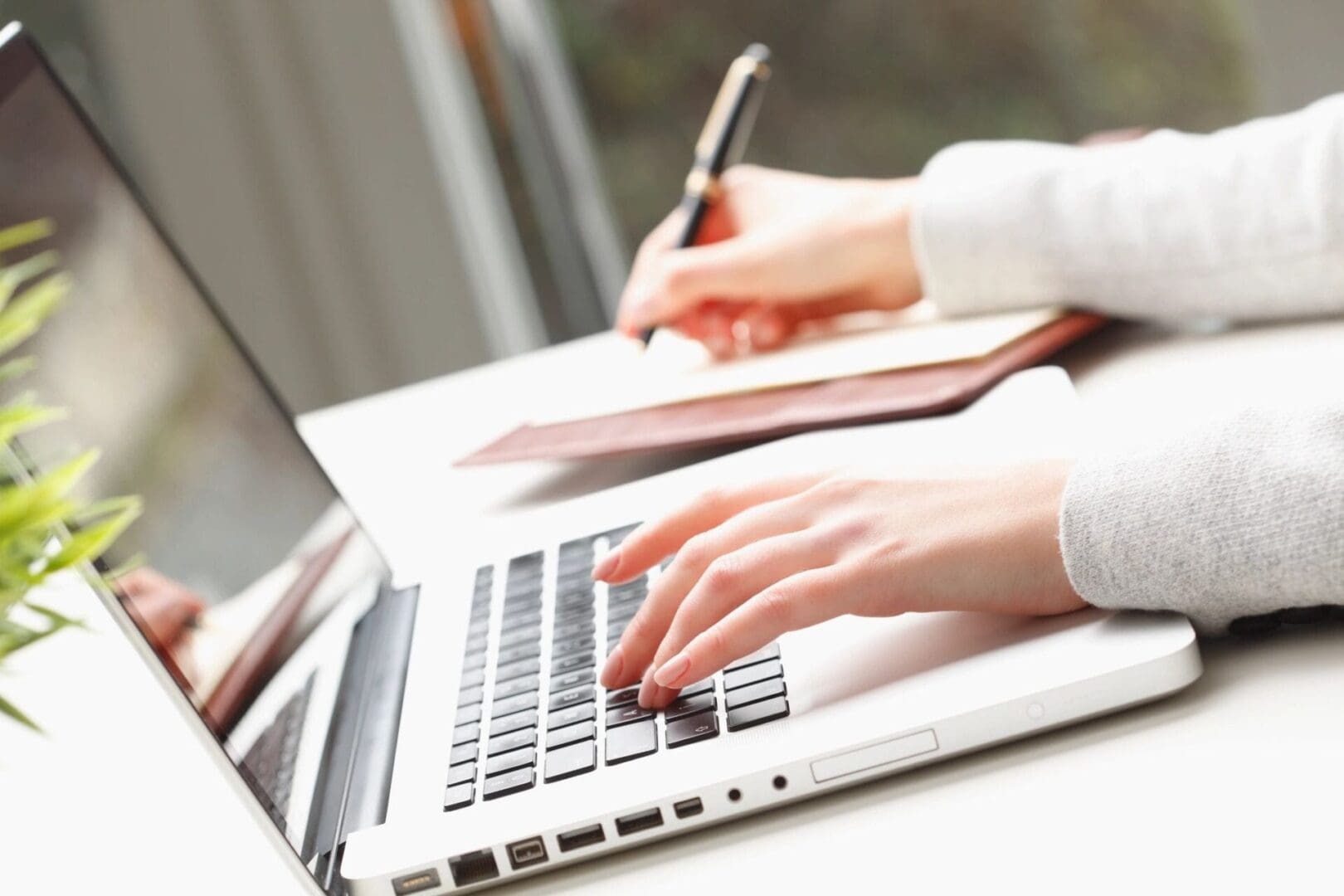 A person typing on a laptop with a pen and paper.