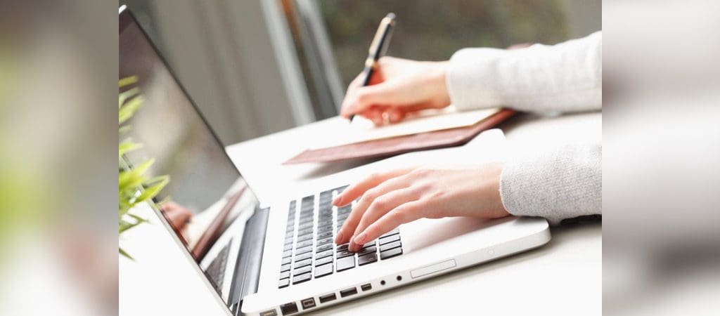 A person typing on a laptop with a pen and paper.